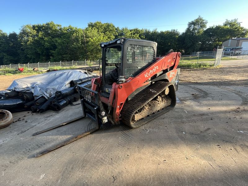 Tracked skid steer with pallet forks beside rolls of black pipe